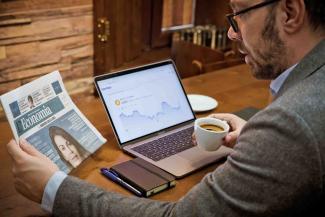 Man reading newspaper next to laptop and phone